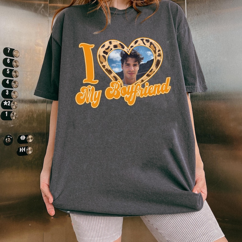 May include: Dark gray t-shirt with the text "I ❤️ My Boyfriend" in orange script. The heart is filled with a leopard print pattern and a photo of a man. The shirt is a casual, oversized style.