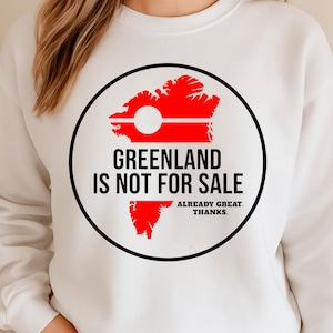 May include: White sweatshirt with a circular graphic. The graphic features a red Greenland outline with a white circle and horizontal lines, and the text "GREENLAND IS NOT FOR SALE" in black. Below it reads "ALREADY GREAT. THANKS."