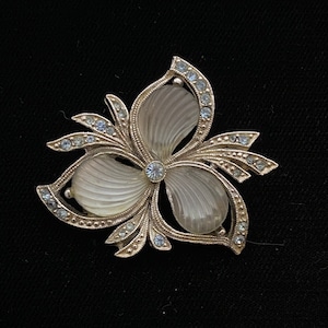 Peut inclure: Une broche en argent avec un motif de fleur blanche. La fleur a trois pétales et est entourée de petits strass.