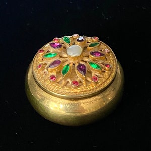 May include: A gold-toned trinket box with a round, domed lid. The lid is decorated with a floral design of green, pink, and purple gemstones surrounding a white pearl.