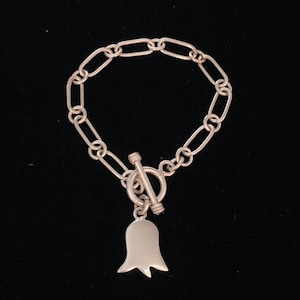 May include: A silver-toned bracelet with an oval link chain and a toggle clasp. A bell-shaped charm hangs from the clasp. The bracelet is displayed against a black background.
