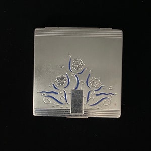 May include: A silver compact with a blue floral design. The compact has a hinged lid and a mirror inside.