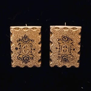 May include: A pair of gold-colored rectangular earrings. Each earring features a textured surface with a dark, ornate design in the center. The design includes floral and cross-like elements.