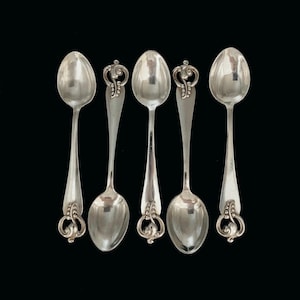 Puede incluir: Cinco cucharas plateadas con mangos ornamentados y cuencos ovalados sobre un fondo negro. Dos cucharas tienen un diseño de mango decorativo, mientras que las otras tres tienen un diseño más simple. Las cucharas son reflectantes y parecen ser de metal.