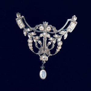 Op de afbeelding: Een zilverkleurige metalen broche met een ingewikkeld opengewerkt ontwerp en heldere strass-steentjes in het frame. Het sieraad heeft een symmetrische, gebogen vorm met bladmotieven en een enkele ovale hanger van iriserende steen.