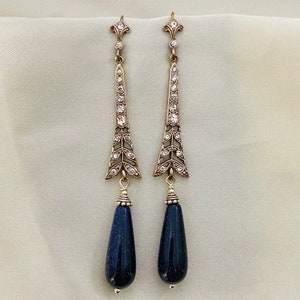 Può includere: Un paio di orecchini pendenti lunghi. Gli orecchini presentano un design traforato decorativo con cristalli trasparenti e una pietra blu scuro a forma di goccia. Il metallo ha un aspetto vintage e antico.