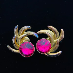 May include: A pair of gold tone earrings with pink crystal stones. The earrings feature a swirling design.