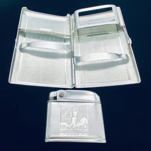 May include: A silver-toned metal cigarette case with an open interior featuring two spring-loaded metal bars and a matching silver-toned metal lighter. The lighter features an embossed graphic of a person playing golf with the word GOLF below it.