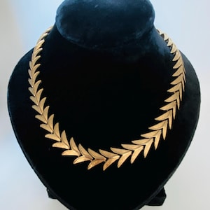 May include: A gold-toned necklace featuring a continuous chain of repeating, textured leaf-shaped links. The metal links are arranged in a symmetrical, chevron-like pattern that forms a circular collar shape, showcasing a matte finish.