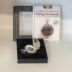May include: Silver-plated Flying Scotsman pocket watch, open to show the clock face, presented in a black box. The watch comes with a certificate of authenticity. The box is on a white surface.