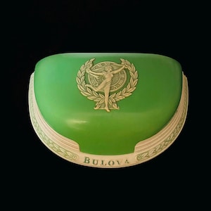 Puede incluir: Una caja de reloj vintage Bulova verde y blanca con una imagen en relieve de una mujer sosteniendo una corona de laurel. La caja tiene una parte superior redondeada y un borde decorativo.