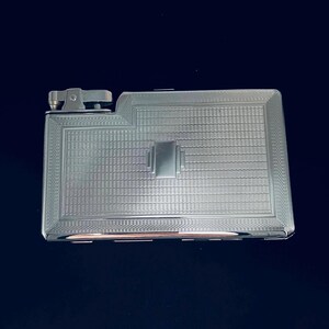 May include: A silver-colored cigarette case with a built-in lighter. The case has a rectangular shape with a textured pattern and a rectangular design in the center. The lighter is attached to the side of the case.