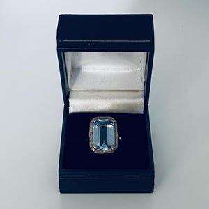 May include: An aquamarine ring with an emerald cut gemstone is displayed in a black jewelry box. The ring has a gold-colored setting with an ornate design around the gemstone. The box is open, revealing the ring against a black velvet interior.