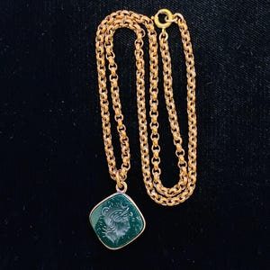 Pode incluir: Um colar de corrente de metal em tom dourado com um pingente de formato quadrado e cantos arredondados. O pingente contém uma pedra verde-escura com o perfil gravado de uma figura clássica voltada para a direita, em uma moldura de metal dourado.