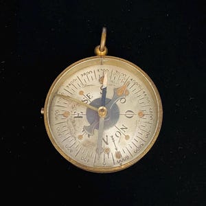 May include: A round brass pocket compass featuring a metallic face with cardinal directions N, S, E, and W, along with numerical markings ranging from 400 to 6000. The device includes a central needle, a small top loop for attachment, and a clear cover.