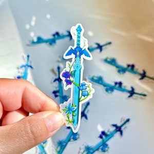 Peut inclure: Plusieurs autocollants repr&eacute;sentant une &eacute;p&eacute;e bleue stylis&eacute;e avec des &eacute;l&eacute;ments floraux. Les autocollants pr&eacute;sentent une lame bleu clair, une garde bleue et sont d&eacute;cor&eacute;s de fleurs bleues et violettes. Fond blanc.