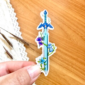 Peut inclure: Un autocollant bleu clair repr&eacute;sentant une &eacute;p&eacute;e stylis&eacute;e avec une garde et une lame bleues. L'&eacute;p&eacute;e est orn&eacute;e de fleurs violettes et bleues et de vignes vertes. L'autocollant est tenu sur une surface en bois.
