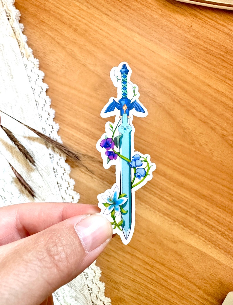 Peut inclure: Un autocollant bleu clair repr&eacute;sentant une &eacute;p&eacute;e stylis&eacute;e avec une garde et une lame bleues. L'&eacute;p&eacute;e est orn&eacute;e de fleurs violettes et bleues et de vignes vertes. L'autocollant est tenu sur une surface en bois.