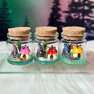Puede incluir: Tres frascos de vidrio con tapas de corcho, cada uno contiene una figura en miniatura de gnomo con una casa de hongo. Los gnomos llevan sombreros de diferentes colores: morado, azul y rojo. Los frascos están llenos de musgo verde y pequeños elementos decorativos.