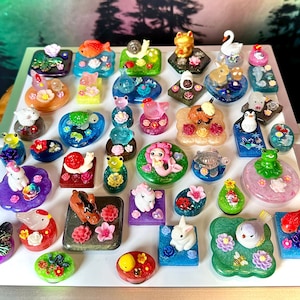 Può includere: Una collezione di piccole statuette in resina colorata raffiguranti vari animali, creature mitiche e insetti, ciascuna montata su una base decorativa ornata con fiori in miniatura, foglie e accenti glitterati di varie forme e dimensioni.
