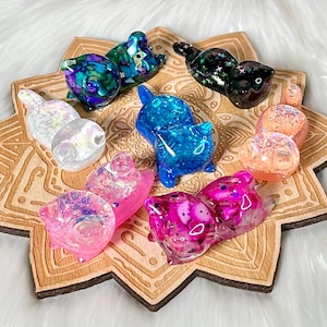 Op de afbeelding: Acht kleine kattenbeeldjes van hars in verschillende kleuren en patronen, waaronder iriserend roze, blauw, groen en zwart, gepresenteerd op een houten dienblad met een mandala-ontwerp.