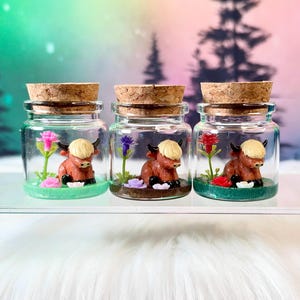 May include: Three clear glass jars with cork stoppers, each containing a miniature brown highland cow figurine, small colorful artificial flowers, and a base of colored glittery material in green, brown, and dark green shades.