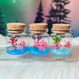 Könnte beinhalten: Drei kleine Glasgefäße mit Korkstopfen, die jeweils eine Miniaturfigur eines rosa Axolotls enthalten. Die Gefäße verfügen über einen blauen Harzboden, kleine künstliche Wasserpflanzen und dekorative Elemente für eine Unterwasserszene.