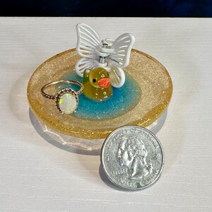 May include: A small, yellow rubber ducky ring holder with a white butterfly on top. The ring holder is filled with blue glitter and has a gold glitter rim. A ring with a white opal stone is sitting on the holder.