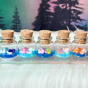 Puede incluir: Cinco pequeñas botellas de vidrio con tapones de corcho, cada una contiene una figurita en miniatura de una criatura marina en agua azul. Las figuritas son de color rosa, morado, azul, naranja y amarillo.