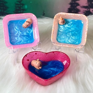 May include: Three resin dishes: two square and one heart-shaped. Each dish has a blue, swirled interior with a small, light brown capybara figurine. The square dishes are pink and peach, while the heart-shaped dish is pink.