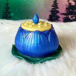 May include: A blue lotus flower-shaped trinket box with a yellow lid and a blue bud on top. The box sits on a green leaf-shaped base. The box has a glittery finish.