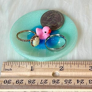 May include: A turquoise blue ring dish with a pink rubber ducky, a silver ring, and a gold ring with an opal stone. The dish is 2 inches in diameter and is next to a ruler for size reference.