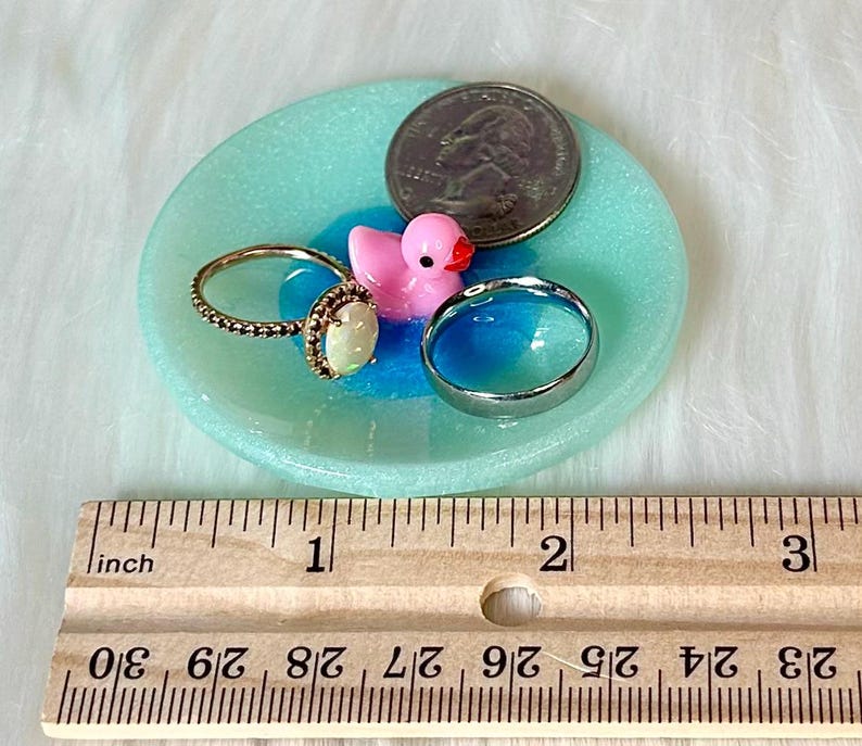May include: A turquoise blue ring dish with a pink rubber ducky, a silver ring, and a gold ring with an opal stone. The dish is 2 inches in diameter and is next to a ruler for size reference.