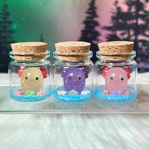 May include: Three glass jars with cork stoppers, each containing a small, colorful creature. The creatures are beige, purple, and pink, with red accents and sit on a blue base with floral details. The jars are displayed on a white surface.