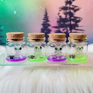 May include: Four small clear glass jars with cork stoppers, each containing a tiny translucent figurine with a distinct facial expression. The jars sit on a clear surface, with purple or green liquid bases and small decorative beads inside.