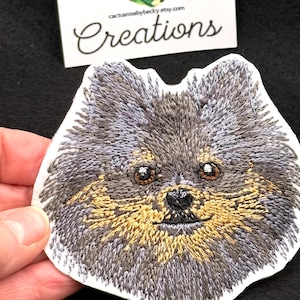 May include: Embroidered patch of a Pomeranian dog's face. The dog has gray and brown fur, with a light brown muzzle and black nose. The patch is on a white background.