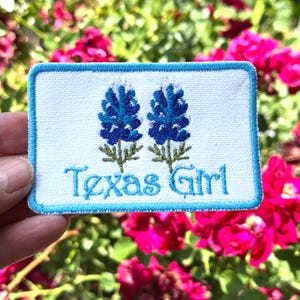 May include: A rectangular patch with a white background and a blue border. The patch features two bluebonnet flower illustrations and the text "Texas Girl" in blue script. The patch is held in front of a background of pink flowers.