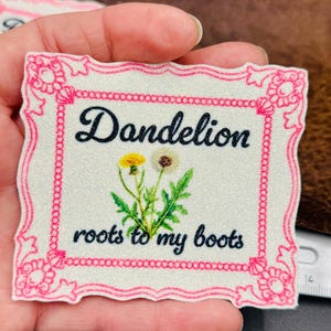 May include: A square patch with a pink floral border and the words "Dandelion roots to my boots" in black script. The patch features a watercolor illustration of a dandelion flower.