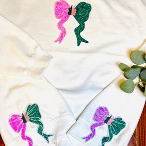 May include: White sweatshirts with glittery bow designs. The bows are split in half, with one side pink and the other green. The design is repeated on the sleeves. A simple, yet eye-catching design for casual wear.