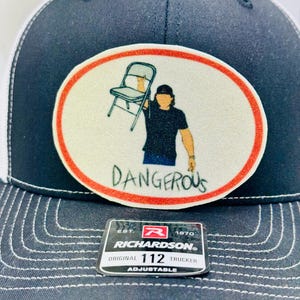 May include: A navy blue and white trucker hat with a patch featuring a person holding a chair, with the word "DANGEROUS" below. The hat has a curved brim and an adjustable strap.