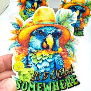 May include: A colorful sticker featuring a blue parrot wearing sunglasses and a yellow hat. The parrot is holding a cocktail and the sticker reads "It's 5 O'Clock SOMEWHERE" in green text. The sticker has a tropical theme.