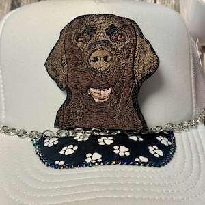 May include: White trucker hat with a brown embroidered dog patch and a black and white paw print trim with rhinestones.