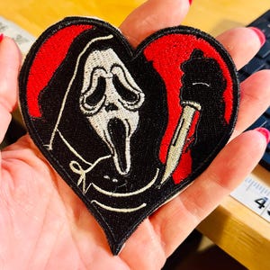May include: Heart-shaped patch featuring the Ghostface mask from the Scream horror film series. The design includes a black background, red accents, and a knife. The patch is likely for clothing or accessories.