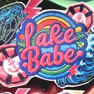 May include: A colorful sticker with the words "Lake Babe" in pink script. The design includes a rainbow, waves, a life preserver with an anchor, and lightning bolts. The sticker has a glittery finish.