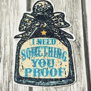 May include: A glittery sticker in the shape of a bottle with a bow. The bow has a leopard print design. The sticker has the text "I NEED SOMETHING YOU PROOF" in blue. A small gold star is in the center.