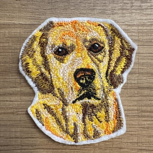 May include: Embroidered patch of a golden retriever dog's head. The dog has brown eyes and a black nose. The patch is on a white background.
