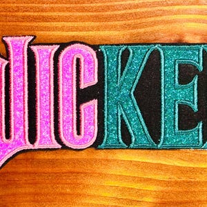 May include: A pink and teal glitter iron-on patch with the word "WICKED" in a bold, stylized font.
