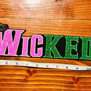 May include: A black felt patch with pink and green glitter letters spelling "WICKED" with a black witch's hat with gold glitter accents on the left side.