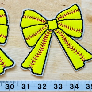 May include: Two yellow and red softball-themed fabric bow patches. The bows are stitched with red stitching and have a yellow background with red stitching.