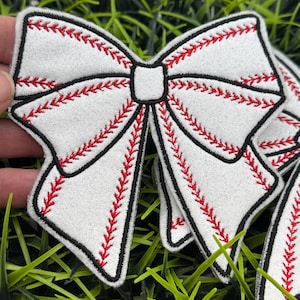 May include: White felt bow with red stitching, resembling a baseball, with black outlining. The bow is on a green grassy surface.